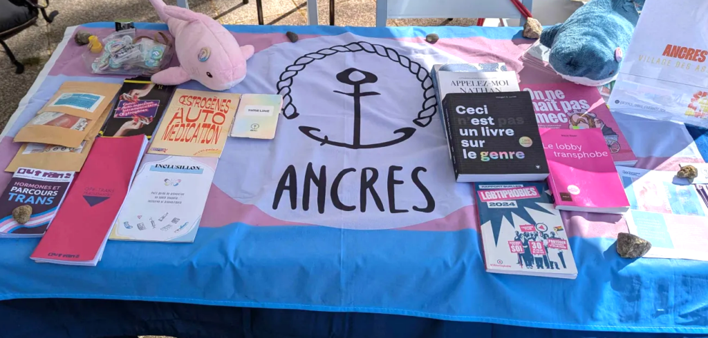 Une photo d'un stand d'ANCRES avec un drapeau trans, des livres et fascicules sur la transidentité